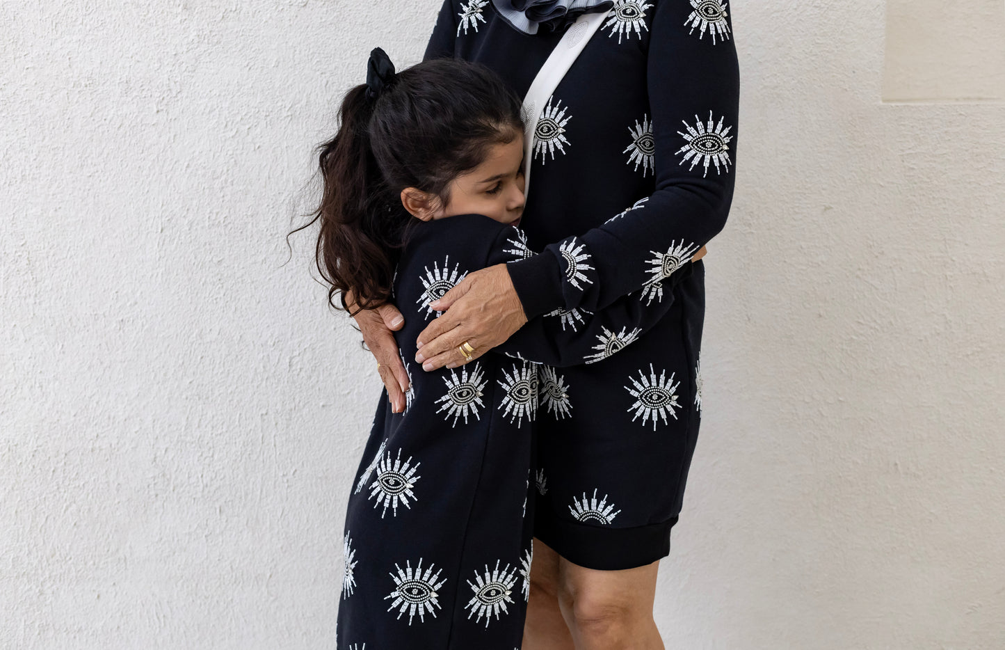 A little girl hugging a woman, both wearing a sustainably and ethically handcrafted black sweatshirt dress with a beaded eye pattern.