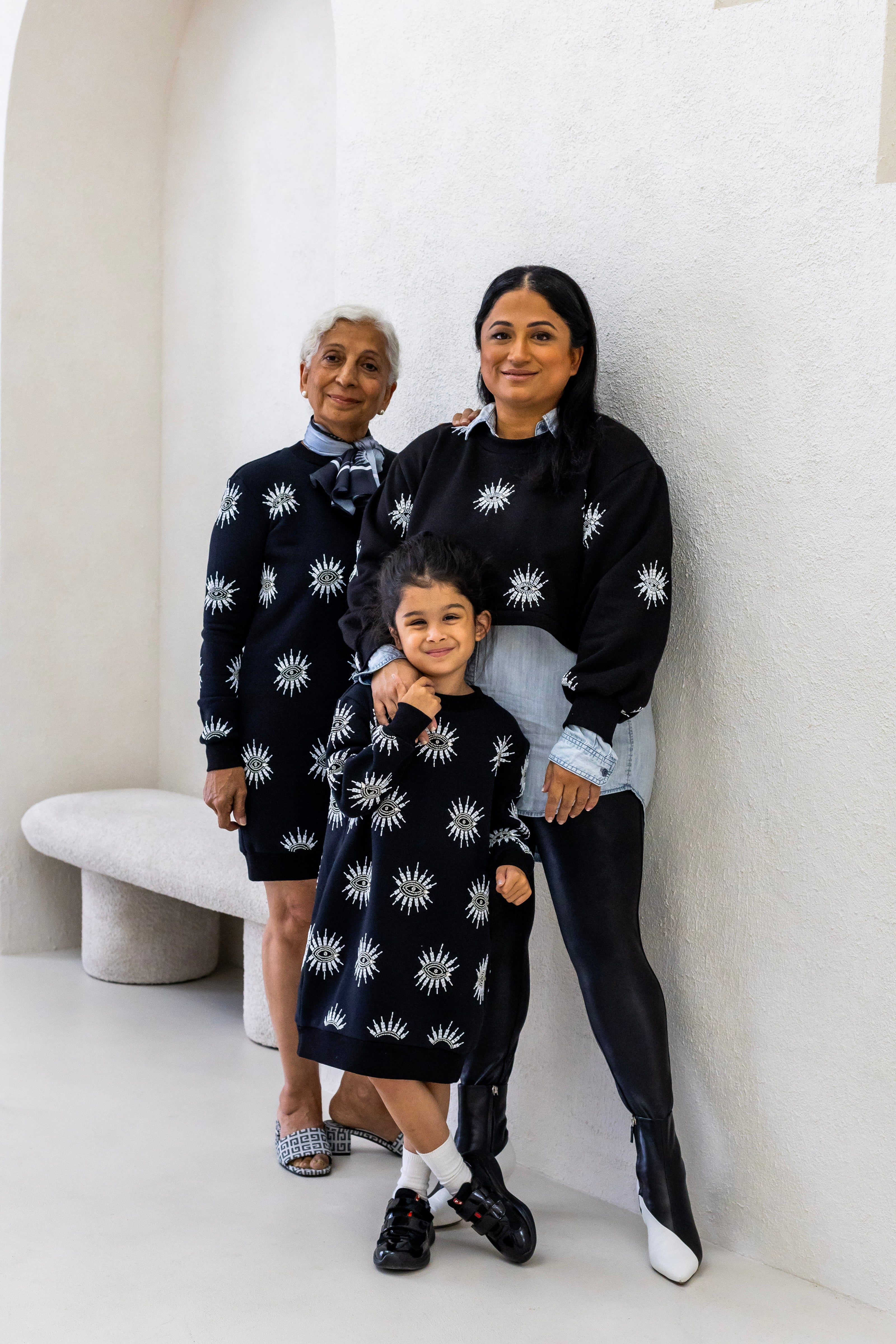 Two women and a little girl wearing sustainably and ethically handcrafted black sweatshirt dresses with a beaded eye pattern.