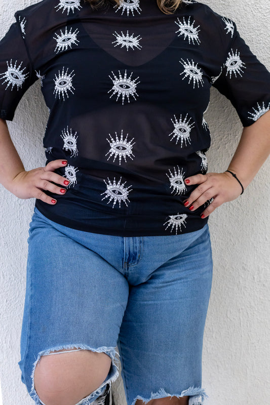 Woman wearing a sustainably and ethically handcrafted sheer black shirt with a white embroidered eye pattern.